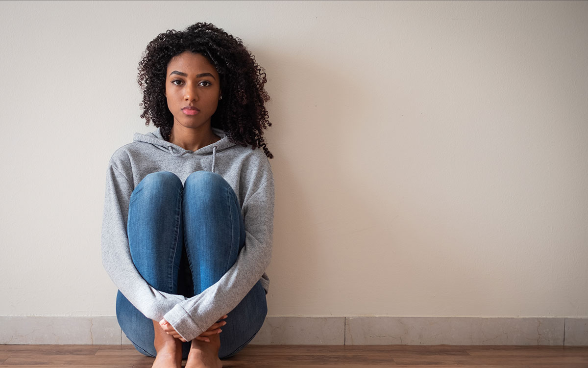 Young african american teenager sitting against a wall, looking sad.