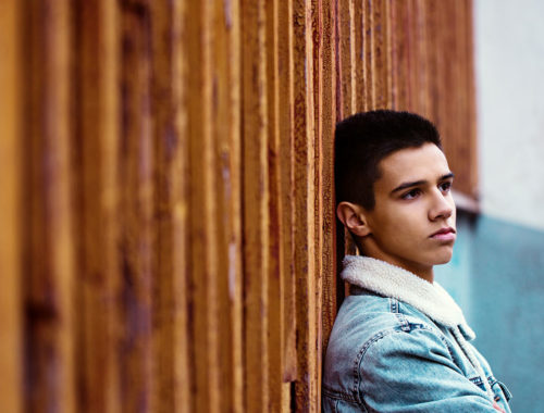 Young latino teen boy sitting up against a wall, looking depressed