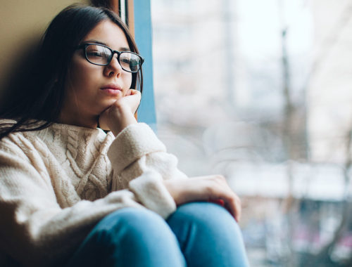 Young latina teen in glasses, sitting by a large window, looking sad and stressed.