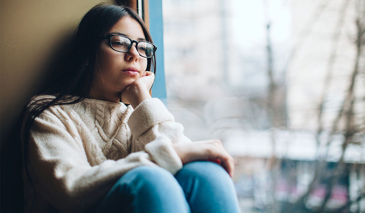 Young latina teen in glasses, sitting by a large window, looking sad and stressed.