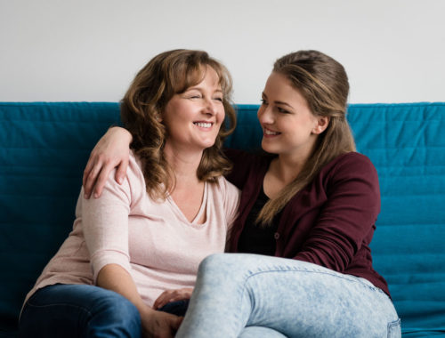 Caucasian mother and daughter sitting on blue couch, with their arms around each other.