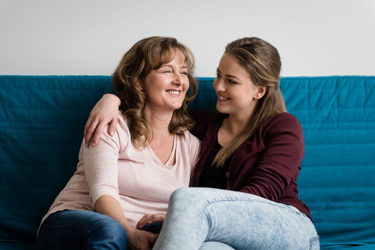 Caucasian mother and daughter sitting on blue couch, with their arms around each other.