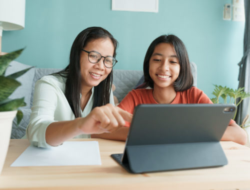 Asian mother smiling and helping teen daughter set intentions on her tablet.