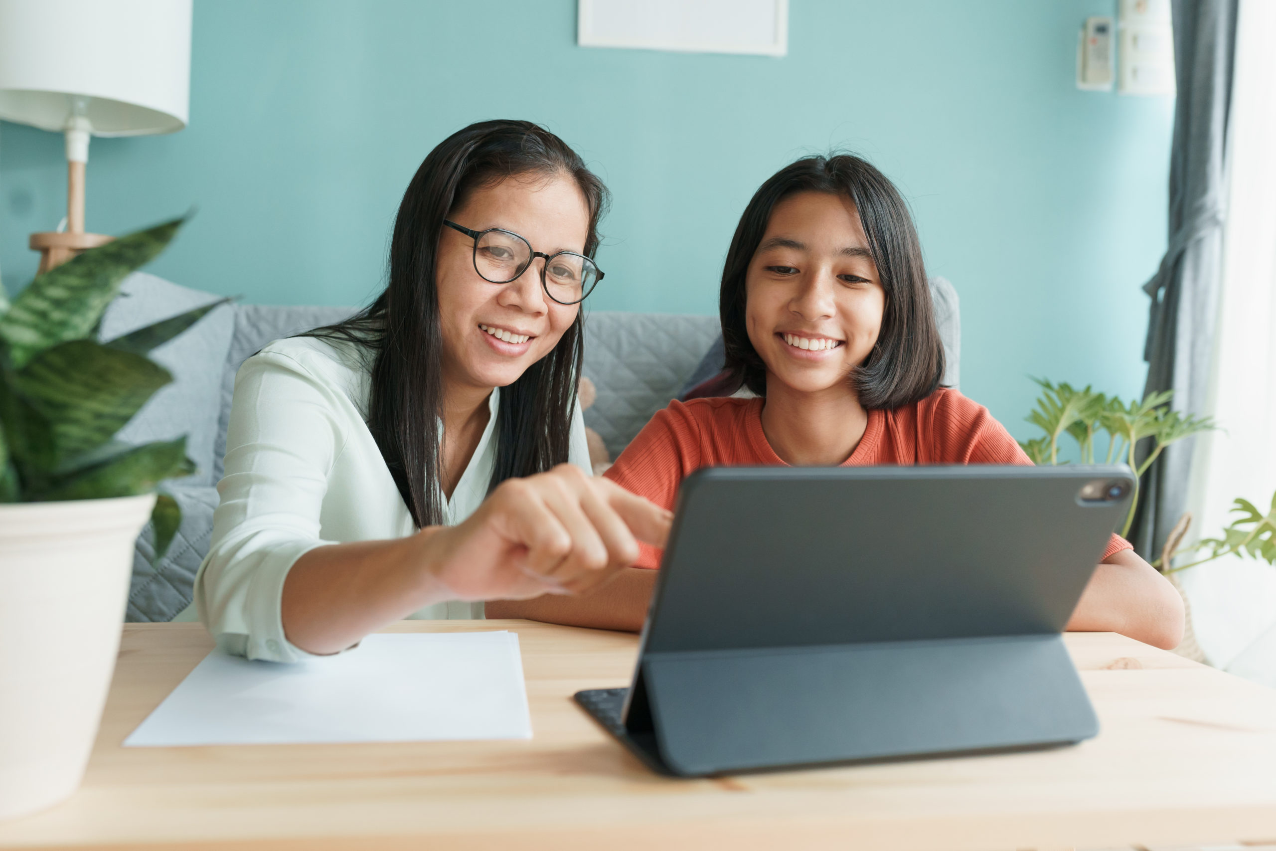Asian mother smiling and helping teen daughter set intentions on her tablet.
