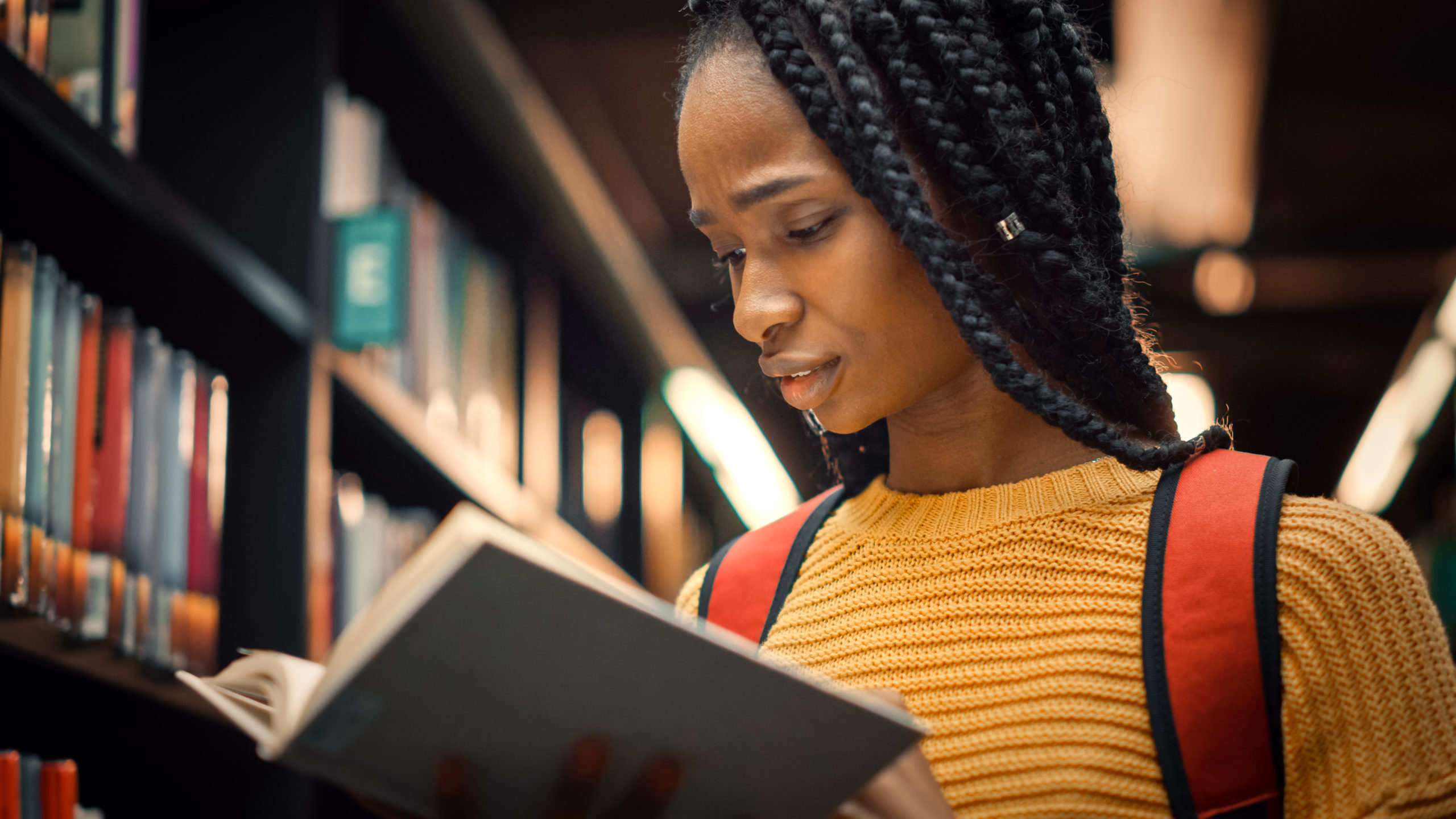 Young black teenager with braids, reading a book in a library.