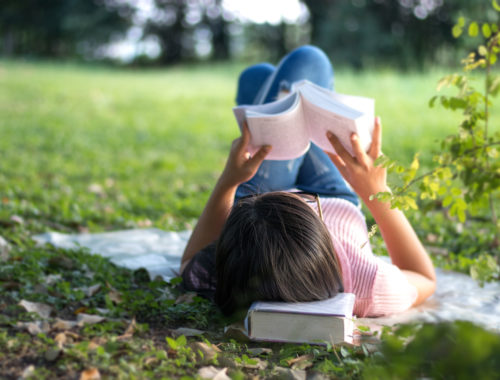 Teen girl reading a book, lying on a picnic blanket in the park.